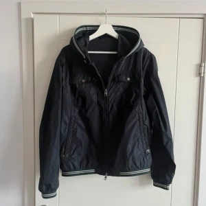 Svart vindjacka från Moncler - Svart vindjacka från Moncler med huva och vita/gröna ränder på muddar och huva. Jackan har två bröstfickor med knappar, två sidofickor med dragkedja och Moncler-logga på ärmen. Perfekt för dig som gillar sportig och clean stil.