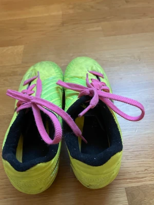 Nike fotbollsskor gul/rosa strl 33.5 - Säljer ett par Nike fotbollsskor i en riktigt cool gul färg med svarta detaljer och rosa snörning. Skorna har rund tå och platt sula med dobbar för gräsplan. Insidan är svart med grönt Nike-tryck. Perfekt för dig som vill sticka ut på planen!
