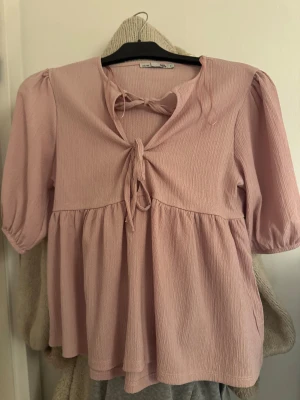 Ljusrosa blus med knyt och puffärm - Supersöt ljusrosa blus från Lager 157 med knyt framtill och lätt puffade ärmar. Blusen har en lös passform.