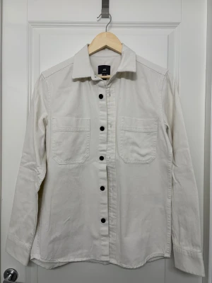 Vit overshirt från H&M med knappar - Snygg vit overshirt från H&M i manchestertyg med svarta knappar framtill och två stora bröstfickor. Skjortan har klassisk krage och lång ärm, perfekt för lager-på-lager och chill stil. Passar dig som gillar enkel och clean look.