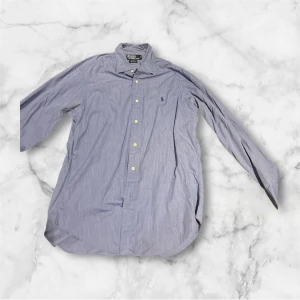 Blå skjorta från Polo Ralph Lauren - Snygg blårandig skjorta från Polo Ralph Lauren med klassisk krage och broderad logga på bröstet. Skjortan har långa ärmar och knappar framtill. En liten missfärgning under armhålan men inte något som syns vid användning. Perfekt för dig som gillar stilrena och tidlösa plagg.