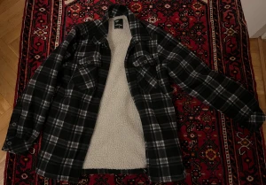 Svart och vit rutig overshirt med teddyfoder - Riktigt snygg svart och vit rutig overshirt med mjukt teddyfoder på insidan. Jackan har två bröstfickor med knappar, klassisk krage och långa ärmar med manschettknappar. Perfekt för lager-på-lager och chill höst- och vinterstil.