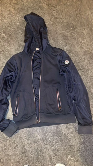 Mörkblå vindjacka från Moncler - Snygg mörkblå vindjacka från Moncler med huva och två framfickor med dragkedja. Jackan har ribbade muddar och Moncler-logga på ärmen. Materialet är lätt och syntetiskt, perfekt för dig som gillar stilren design och sportig vibe.