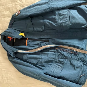 Blå vindjacka från Parajumpers L - Säljer en blå vindjacka från Parajumpers i storlek L. Jackan har flera praktiska fickor, coolt patch på ärmen och detaljer i gult och orange. Materialet är lätt polyester och jackan har dragkedja framtill. Perfekt för dig som gillar street och funktion. Säljer den för att den inte passar mig längre 