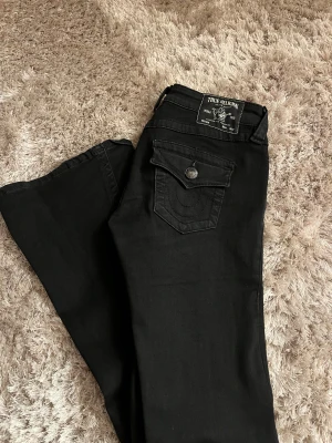 Svarta bootcut jeans True Religion - Svarta low waist jeans från True Religion med bootcut passform och klassisk femficksdesign. Snygga detaljer på bakfickan med lock och knapp, samt tydlig logotyp på bakre linningen. Jeansen har en stilren look och är tillverkade i ett mjukt jeansmaterial. 