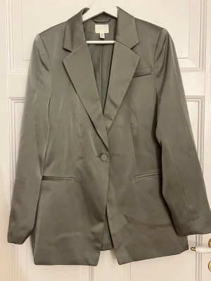 Grågrön kavaj från H&M i satinlook - Stilren grå kavaj från H&M i storlek M. Kavajen har en snygg satinlik finish, klassisk krage, en knapp framtill och raka fickor. Perfekt för dig som gillar en clean och modern look. Passar dig som vill ha en enkel men elegant kavaj.