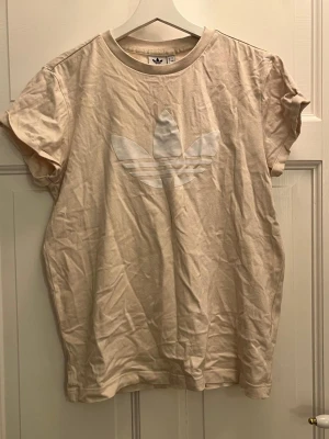 Beige Adidas t-shirt med logga - Snygg beige t-shirt från Adidas med stor vit Trefoil-logga på bröstet. Klassisk rund halsringning och korta ärmar. Tillverkad i mjuk bomull för en skön känsla. Perfekt för en avslappnad och sportig stil.