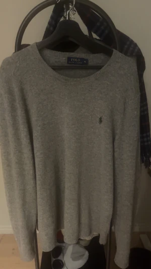 Grå stickad tröja Polo Ralph Lauren XL - Snygg grå stickad tröja från Polo Ralph Lauren i storlek XL. Tröjan har lång ärm, rund hals och den klassiska broderade loggan på bröstet. Perfekt för dig som gillar enkel och stilren design med premiumkänsla.