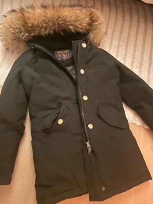 Svart parkas Woolrich med päls - Svart parkas från Woolrich med fluffig päls på huvan, snygga knappar och dragkedja framtill. Jackan har två fickor med lock och är perfekt för kalla dagar. Klassisk och stilren modell som är lite längre i passformen. Säljer eftersom den är för stor på mig, priset kan diskuteras