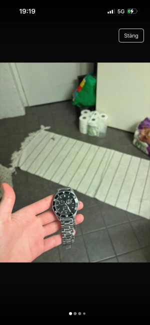 Silverfärgad Everone klocka - Stilren klocka från Everone med svart urtavla och tre mindre subdials. Boetten och länken är i silverfärgat metall, vilket ger en klassisk och clean look. Perfekt accessoar för dig som gillar en sportig men ändå tidlös stil.