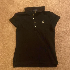 Svart kortarmadpiké från Polo Ralph Lauren - Svart pikétröja från Polo Ralph Lauren med klassisk krage och korta ärmar. Tröjan har tre vita knappar framtill och den ikoniska broderade loggan i vitt på bröstet. Tillverkad i mjuk bomull som är skön mot huden och har en slim passform.