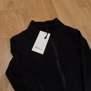 Svart Define Jacket från Lululemon - Svart define jacket från Lululemon med hel dragkedja framtill och hög krage. Jackan är tillverkad i ett lätt syntetmaterial vilket gör den smidig och perfekt för aktiva dagar. Stilren design utan mönster och diskreta sömmar. Liten logga på dragkedjorna samt en på ryggen strax under nacken. Skickas självklart med originalförpackning!