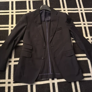 Svart kavaj från Rose & Born - Stilren svart kavaj från Rose & Born, tillverkad i Italien. Klassisk modell med två knappar framtill, flera fickor med lock och slits baktill. Fodrad insida i mörk nyans. Storleken skulle beskrivas som en liten M. Perfekt för dig som vill ha en tidlös och elegant look.