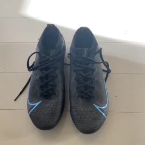 Nike svarta fotbollsskor med blå swoosh - Säljer ett par svarta Nike fotbollsskor! Riktigt bra skick! Slite modellen med konstgräsdobbar. Används en gång en rätt kort stund! Pris kan diskuteras 
