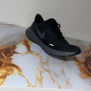 Svarta Nike React sneakers - Snygga svarta Nike React sneakers med mesh-ovandel och mörkgrå sula. Skorna har en sportig design med rund tå, platt sula och klassisk snörning. Perfekta för dig som gillar stilrena och bekväma träningsskor med diskret Nike-logga på sidan.Skorna är helt nya har använts en gång under inomhus sport.
