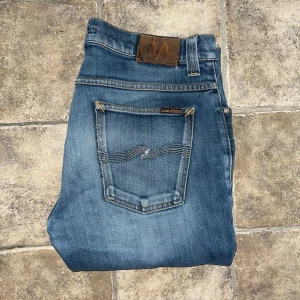 Nudie Jeans Grim Tim  - Snygga blå jeans från Nudie Jeans i modell Gril Tim. Jeansen har slim fit-passning, normal midja och är tillverkade i mjukt jeansmaterial. Midja 42cm, längd 105cm. JAG SKICKAR EJ FLER BILDER NÄR JAG BÄR JEANSEN! för storleksguide hänvisar jag till måtten och för bättre inblick i passformen rekommenderar jag att googla på modellnamnet. Jeansen kan vara uppsydda utan att jag noterat det så kolla måtten noggrant :) OBS se defekt på sista bilden. S4