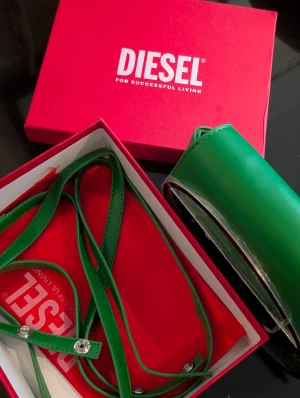 Grön axelväska från Diesel - Snygg grön axelväska från Diesel med stort silverfärgat D-spänne framtill. Väskan har en stilren, rundad form och justerbar axelrem. Tillverkad i slätt skinnliknande material och har magnetlås samt innerfack med dragkedja. Perfekt för dig som vill sticka ut med färg.