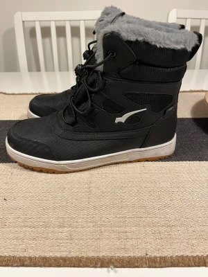 Svarta vinterstövlar från Bagheera - Säljer ett par svarta vinterstövlar från Bagheera med grått fuskpälsfoder och grov mönstrad sula. Skorna har snörning med snabblås, platt sula och rund tå. Ovandelen är i slitstark syntet och textil, perfekt för kalla dagar. Vattenavvisande varmfodrad stövel med velcro för riktigt kalla dagar. Size 40. 25.5 cm Använd bara en gång.