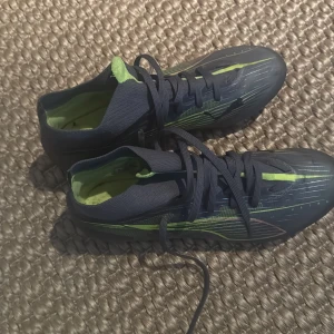 Svarta och limegröna Puma fotbollsskor - Säljer ett par snygga fotbollsskor från Puma i svart med limegröna detaljer och mönster. Skorna har snörning och dobbar för bra grepp på planen. Ovandelen är i syntetmaterial och insidan är fodrad i limegrönt. Perfekta för dig som vill sticka ut på fotbollsplanen.