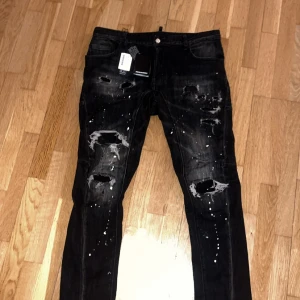 Dsquared2 Slim fit Svart - Säljer ett par svarta skinny jeans från Dsquared2 med slitna detaljer, hål och vit färgstänk på både fram- och baksida. Jeansen har klassisk femficksdesign och är tillverkade i bomull med stretch. Perfekt för dig som gillar en edgy och trendig look. Tags finns.