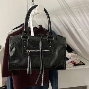 Svart handväska med dragkedja - Cool svart handväska i skinnimitation med snygga detaljer och silverfärgad dragkedja framtill. Väskan har två rejäla handtag, dekorativa nitar och långa remmar som ger en edgy vibe. Perfekt storlek för att få plats med allt viktigt och passar till många olika stilar.