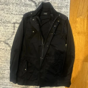 Svart field jacket från Diesel - Svart parkajacka från Diesel med flera fickor framtill, snygga knappar och dragkedja. Jackan har en klassisk krage och detaljer på axlarna. Perfekt för dig som gillar stilrena och funktionella jackor med coola detaljer.