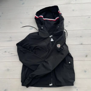 Svart vindjacka från Moncler - Svart vindjacka från Moncler med huva och coola rödvita detaljer på insidan av huvan. Jackan har dragkedja framtill, snörning vid huvan och Moncler-logga på ärmen. Materialet är lätt och syntetiskt, perfekt för blåsiga dagar.