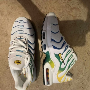 Nike Air Max Plus vita med färgdetaljer - Nike Air Max Plus sneakers i vitt med blå, gröna och gula detaljer. Skorna har synlig Air Max-dämpning, vågformade linjer och svart Swoosh på sidan. Ovandelen är i mesh och syntet, med snörning och platt sula. Perfekta för dig som gillar färgstarka och sportiga sneakers.