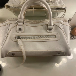 Vit handväska med dragkedja - Stilren vit handväska i skinnimitation med silverfärgade metalldetaljer och praktiskt handtag. Väskan har en dragkedja framtill och rymligt huvudfack. Perfekt för dig som gillar en clean och klassisk look. Köpt i Frankrike 