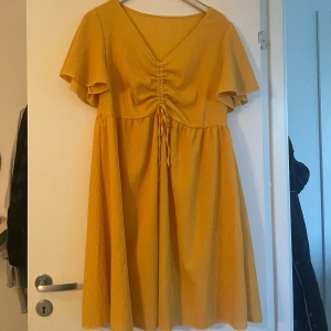 Gul kortklänning med snörning - Superfin gul klänning med v-ringning och snörning framtill som ger en snygg rynkad effekt. Klänningen har korta, fladdriga ärmar och ett lätt, glittrigt tyg som känns som polyester. Passformen är loose och kjolen är utställd för en skön vibe.