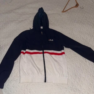 Blå och vit vindjacka från FILA - Snygg vindjacka från FILA i marinblått och vitt med röd detalj över bröstet. Jackan har huva med snörning, hel dragkedja och elastisk nederkant. Perfekt för dig som gillar sportig stil och vill ha något lätt och bekvämt.