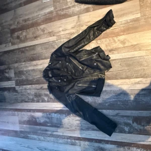 Svart skinnjacka från Saints de Matilde - Svart skinnjacka från Saints de Matilde i klassisk bikerstil med flera dragkedjor, knappar och ett bälte vid midjan. Jackan har långärmade ärmar och coola detaljer som ger en edgy vibe. Perfekt för dig som gillar street och rockig stil.