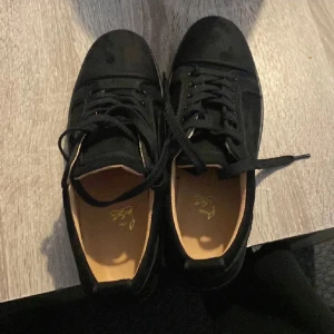 Svarta sneakers från Christian Louboutin - Snygga svarta sneakers från Christian Louboutin med klassisk röd sula och diskret logga. Skorna har rund tå, platt sula och är tillverkade i mocka med svarta snören. Perfekt för dig som vill ha en lyxig och stilren look.                            Det ända dåliga med skorna är att en knapp på sidan av skorna har ramlat av men inget man tänker på 