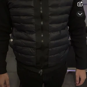 Svart Moncler cardigan alla storlekar - Svart pufferjacka från Moncler med huva och ribbstickade ärmar. Jackan har quiltad framsida, dragkedja och en ficka med zip på ärmen. Moncler-logga på vänster ärm och coolt serietryck på insidan. Perfekt för dig som gillar street och exklusiva märken.