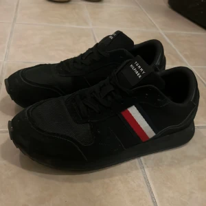 Svarta sneakers Tommy Hilfiger - Snygga svarta sneakers från Tommy Hilfiger med klassisk snörning och rund tå. Skorna har en cool detalj med röd, vit och blå rand på sidan och är tillverkade i en mix av syntet och textil. Perfekta för dig som gillar stilrena och sportiga skor.