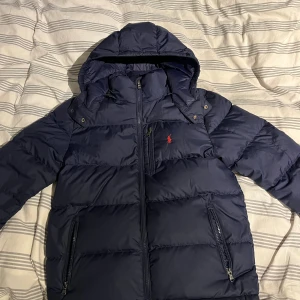 Mörkblå pufferjacka från Polo Ralph Lauren - Snygg mörkblå pufferjacka från Polo Ralph Lauren med huva och dragkedja framtill. Jackan har två sidofickor med dragkedja och en bröstficka, samt Polo-logga i rött på bröstet. Perfekt för kalla dagar och har en clean, sportig vibe. Storlek junior Large (14-16år) 