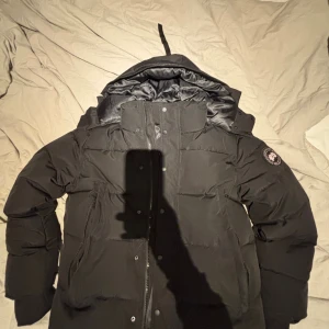 Svart pufferjacka från Canada Goose - Svart pufferjacka från Canada Goose med ikonisk Arctic Program patch på ärmen. Jackan har huva, dragkedja och knappar framtill, samt quiltad design som ger extra värme. Perfekt för kalla vinterdagar och har en clean, stilren look. Päls tillkommer!