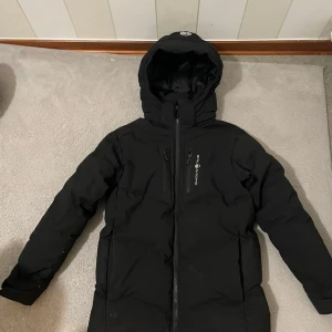 Svart Sail Racing dunjacka med huva - Svart dunjacka från Sail Racing med stor, fodrad huva och flera praktiska fickor med dragkedja. Jackan har en clean look, lång ärm och tydlig logga på bröstet. Perfekt för kalla dagar och har en snygg, sportig vibe.