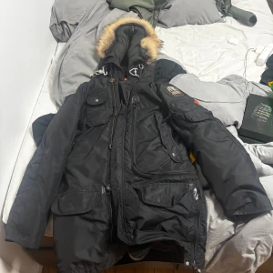 Svart Parajumpers parkajacka med päls - Svart parkajacka från Parajumpers med snygg päls på huvan, flera fickor och coola detaljer som patch på ärmen och gul rem. Jackan har dragkedja och knappar framtill, perfekt för kalla dagar. Klassisk look med modern touch. Byttaa!!!