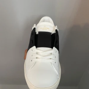 Valentino Garavani Open sneakers - Snygga Valentino Garavani Open sneakers i vitt skinn med en bred svart läderpanel över mitten. Klassisk rund tå, platt sula och vita snören. Bakpartiet har diskreta nitar för extra edge. Perfekt för dig som gillar stilrena och lyxiga detaljer.