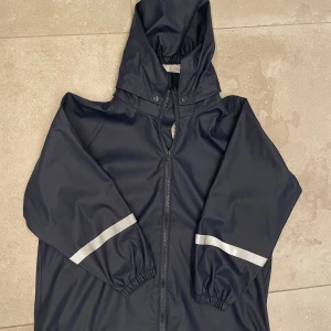 Mörkblå regnjacka med huva Abeko - Mörkblå regnjacka för barn med avtal huva och dragkedja framtill. Jackan har resårmuddar och reflex. 