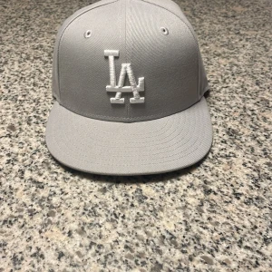 Grå LA Dodgers keps från New Era 55.6 - Snygg grå LA Dodgers keps från New Era 59FIFTY med vit broderad LA-logga framtill och New Era-logga på sidan. Klassisk flat brim och strukturerad form. Tillverkad i bomull och polyester, perfekt för dig som gillar streetstyle och sportig look. Storlek 55.8