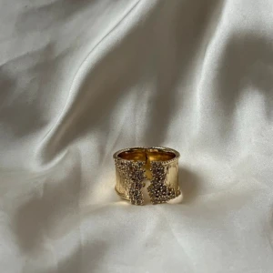 Unik Guldring med vintage look  - Unik bred ring i guldton med öppning framtill och glittriga stenar som dekoration. Ringen har en räfflad yta och detaljerade kanter som ger den en cool och lyxig vibe. Perfekt för dig som vill sticka ut med ett statement-smycke.