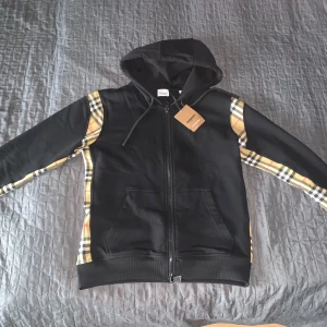 Svart Burberry hoodie med rutiga detaljer - Svart hoodie från Burberry med klassiska beige-rutiga paneler längs ärmar och sidor. Tröjan har huva med snörning, hel dragkedja framtill och två fickor. Perfekt för dig som vill ha en streetig och lyxig look med ikoniska Burberry-vibbar.