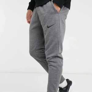 Grå Nike Dri-Fit mjukisbyxor L - Snygga grå mjukisbyxor från Nike med Dri-Fit-teknologi. Byxorna har  elastisk midja med snörning och sidofickor med meshfoder. Nike-logga på benet och ribbade muddar vid benslut. Perfekta för chill eller träning.