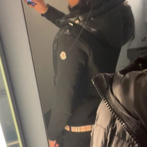 Moncler cardigan  - Svart cardigan från Moncler med glansig finish och huva. Västen har dragkedja framtill, två sidofickor med dragkedja och Moncler-logga på bröstet.