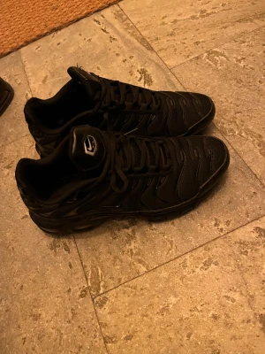 Svarta Nike Tn - Säljer ett par svarta Nike Tn med snyggt vågigt mönster på ovansidan och diskret Swoosh på sidan. Skorna har snörning och är tillverkade i syntet och mesh för bra komfort och andning. Perfekt för dig som gillar stilrena och sportiga skor. Dom är i jätte bra skick säljer för har bytt stil 