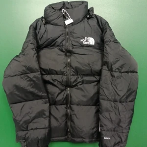 Svart pufferjacka från The North Face - Svart pufferjacka från The North Face med hög krage och tydlig logga på bröstet. Jackan har quiltad design, dragkedja framtill och är tillverkad i syntetmaterial. Perfekt för kalla dagar och har en clean, urban look.