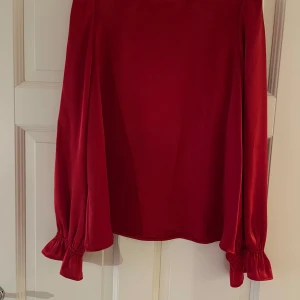 Röd blus med volangärmar - Säljer en röd blus med långa ärmar och volangdetaljer vid ärmsluten. Blusen har en rak passform och är tillverkad i ett glansigt material som ger en lyxig känsla. Perfekt för dig som vill sticka ut med färg och stil.