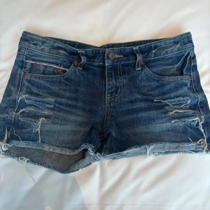 Blå jeansshorts med slitningar - Säljer ett par blå jeansshorts med rå kant och slitna detaljer framtill. Shortsen har klassisk femficksdesign, röda nitar och knappstängning. Perfekt för dig som gillar en avslappnad och edgy stil. Materialet är robust denim och passformen är normal.